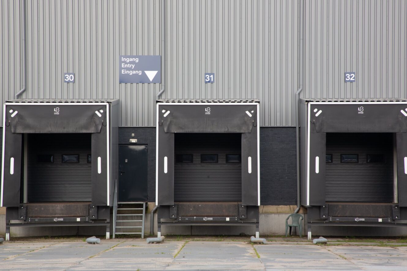 Drie laaddockdeuren genummerd 30, 31 en 32 op een grijs industrieel gebouw, met een bord erboven dat Entry aangeeft in meerdere talen.