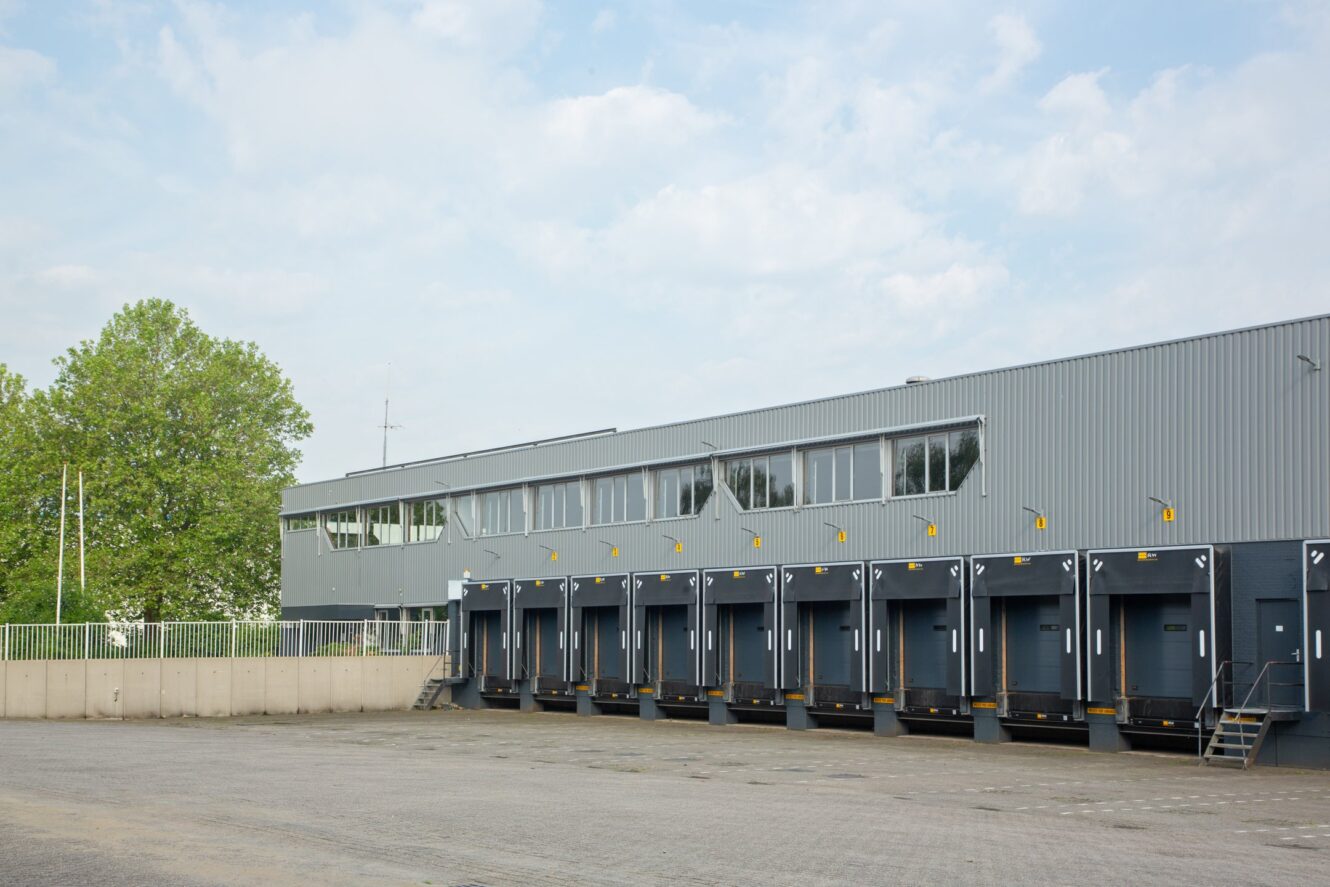 Een groot industrieel pakhuis met meerdere laadkades en een leeg trottoir aan de voorkant, onder een gedeeltelijk bewolkte hemel.