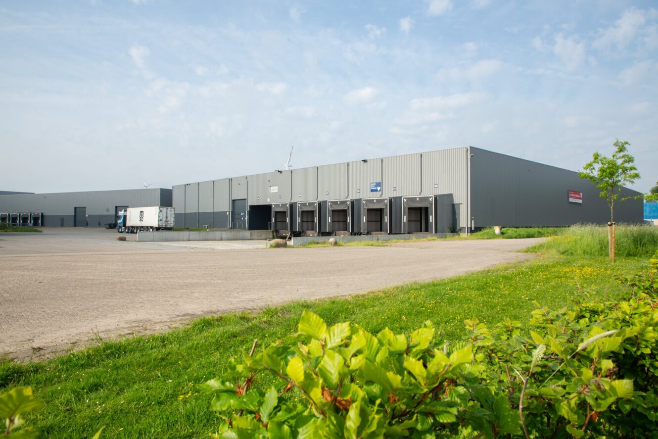 Een groot grijs industrieel pakhuis met verschillende laadkades en een geparkeerde vrachtwagenoplegger, omgeven door een verhard terrein en groen gras met struiken op de voorgrond.