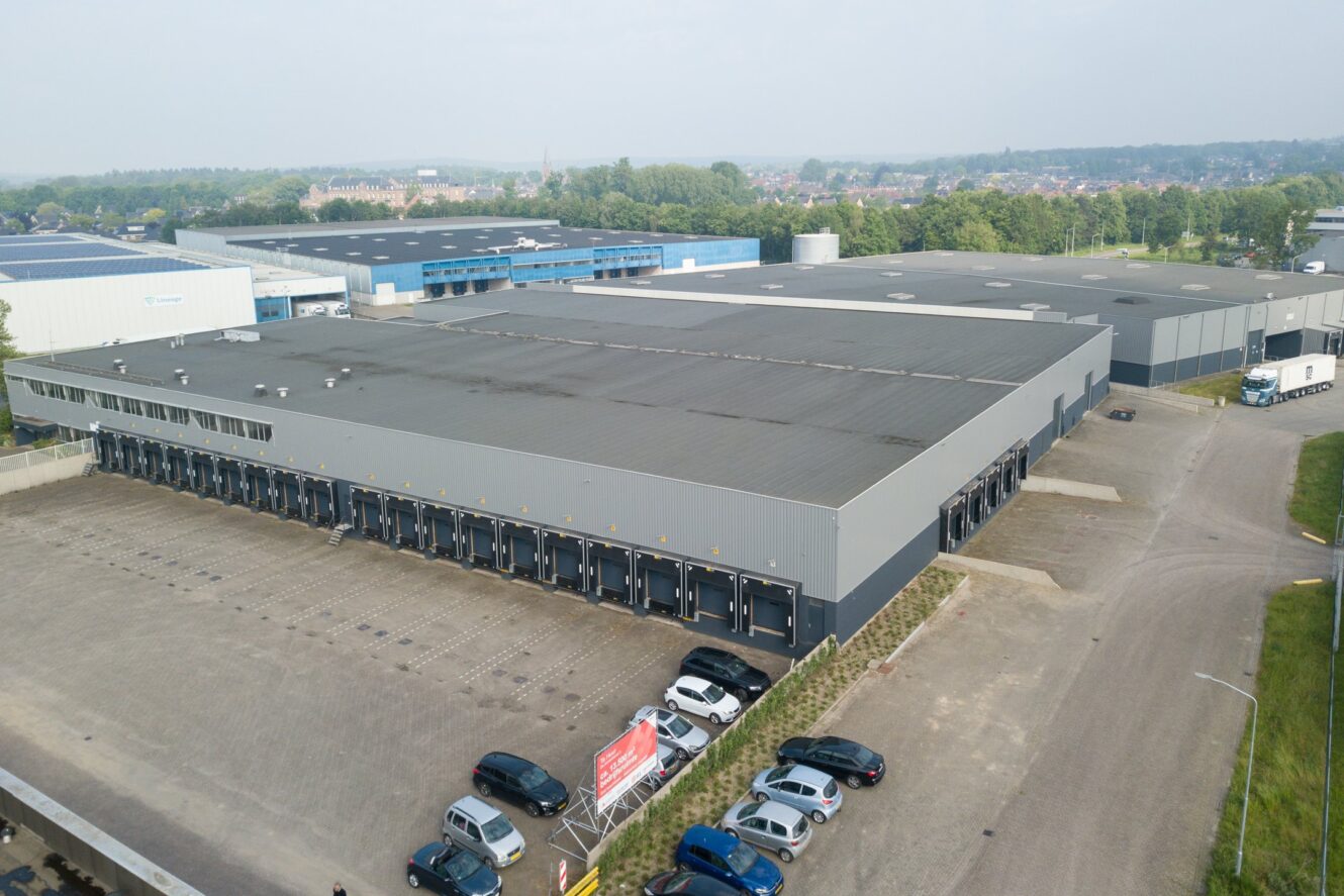 Groot industrieel magazijn met meerdere laaddocks en een parkeerplaats met meerdere auto's, omringd door andere industriële gebouwen en groen op de achtergrond.