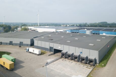 Luchtfoto van een groot industrieel magazijn met laadkades, geparkeerde vrachtwagens en zeecontainers in een bedrijvenpark.