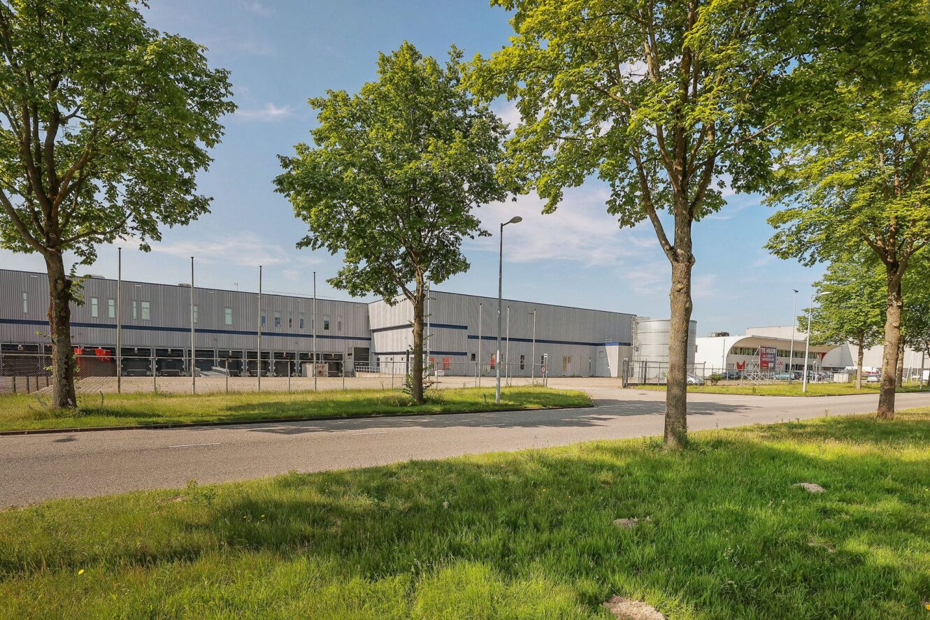 Een groot industrieel gebouw met een blauwe en witte buitenkant is te zien achter een rij bomen en een grasveld langs een weg op een zonnige dag.