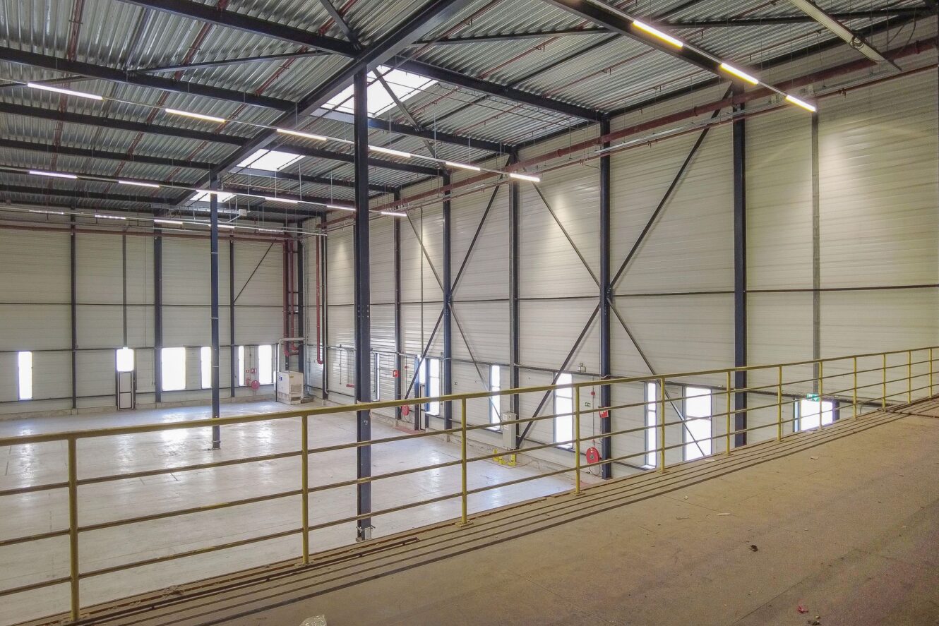 Binnenzicht van een groot leeg pakhuis met hoge plafonds, metalen balken, dakramen en een gele veiligheidsreling langs een verhoogd platform.