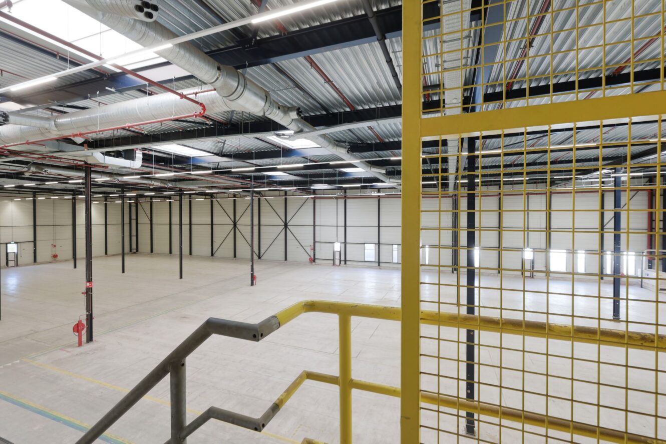 Interieur van een groot, leeg industrieel pakhuis met hoge plafonds, zichtbare leidingen, steunkolommen en een gele metalen trap op de voorgrond.