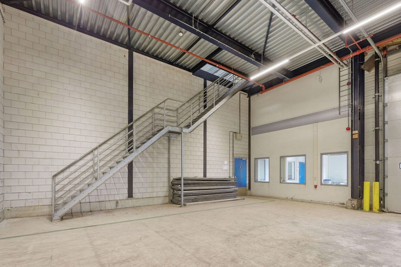 Interieur van een industrieel magazijn met betonnen muren, metalen trap, gestapelde panelen en grote ramen bij een blauwe deur. Bovengrondse leidingen en zichtbare plafondstructuur zijn zichtbaar.