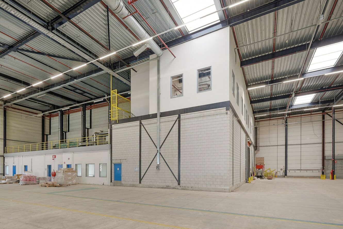 Interieur van een groot leeg pakhuis met hoge plafonds, dakramen en een kantoorgebouw van twee verdiepingen. Langs de muren staan pallets en dozen opgestapeld.