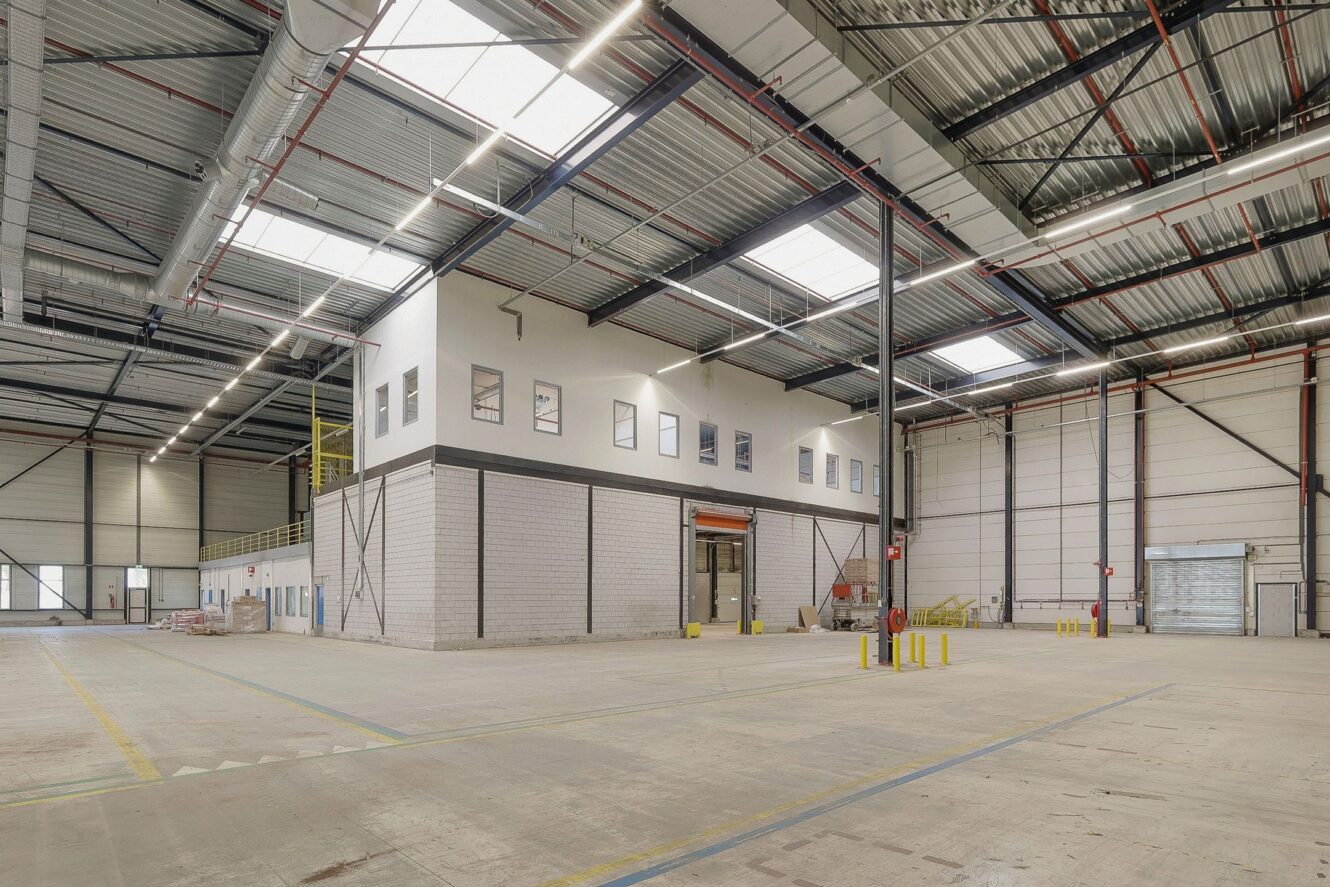 Interieur van een groot, leegstaand industrieel pakhuis met hoge plafonds, zichtbare balken, dakramen en een verhoogd kantoorgedeelte met ramen.