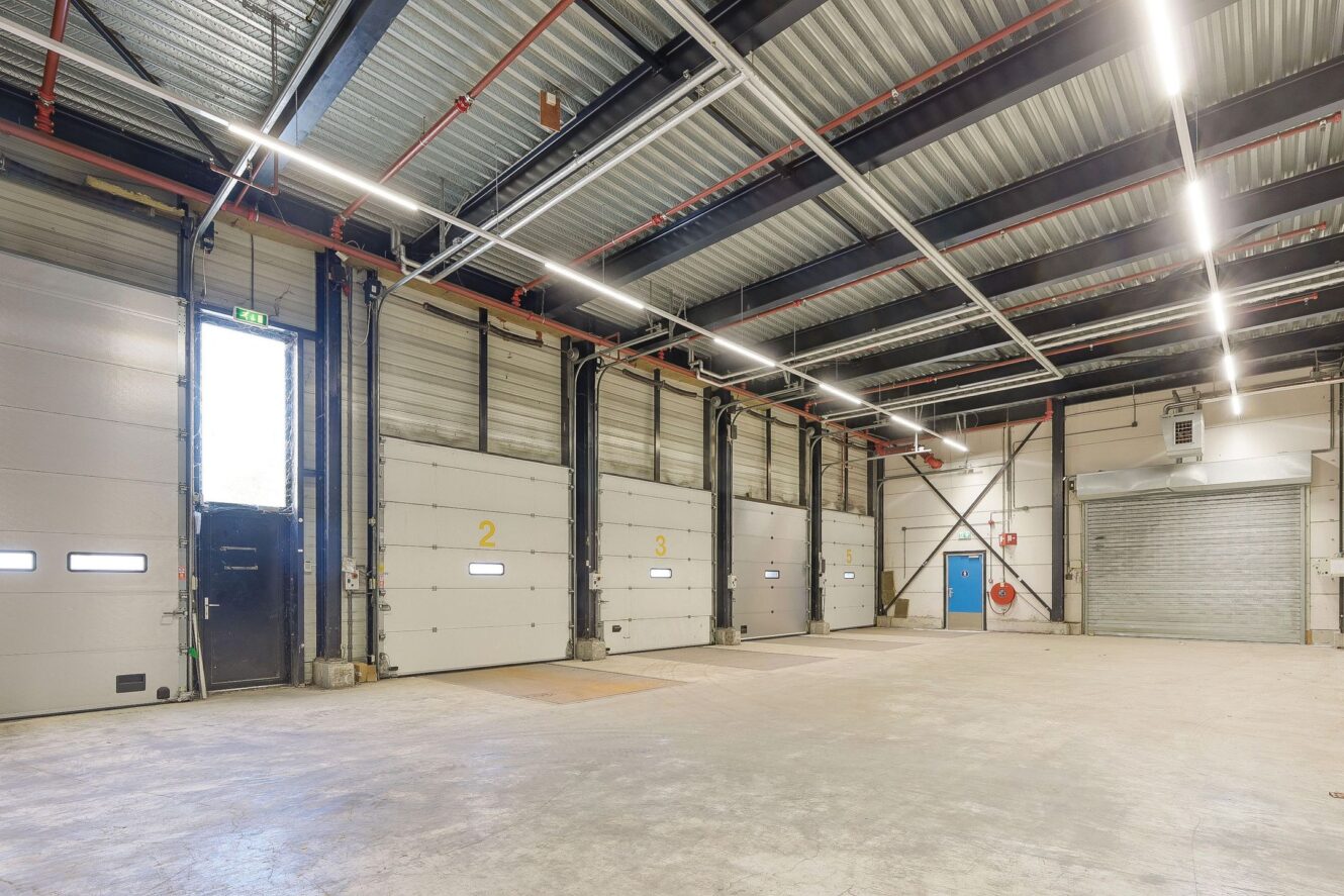 Interieur van een leeg magazijn met hoge plafonds, vijf grote laadperrondeuren genummerd van 1 tot 5 en fluorescerende verlichting boven het hoofd.