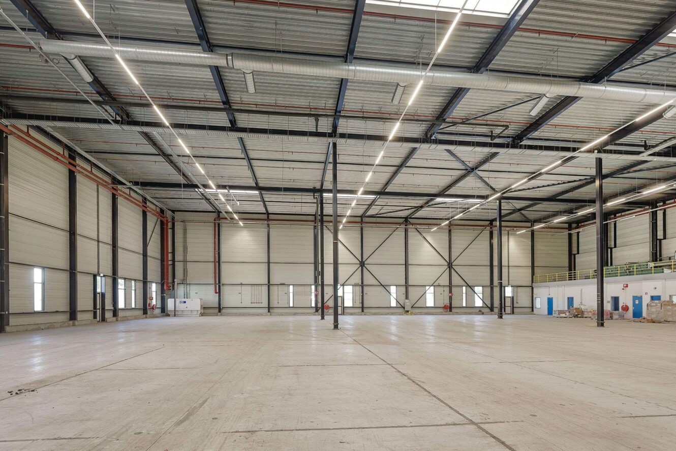 Ruim, leeg industrieel pakhuis met hoge plafonds, betonnen vloeren, metalen steunbalken en tl-verlichting boven het hoofd.