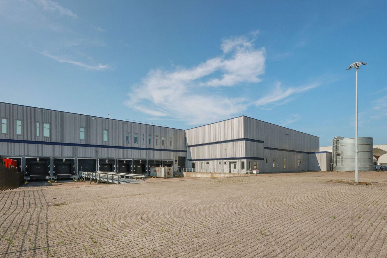 Een groot industrieel magazijn met meerdere laadkades en een breed, leeg geplaveid terrein onder een strakblauwe hemel.