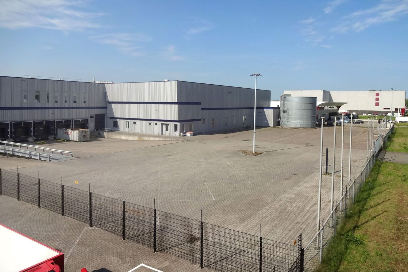 Groot industrieel magazijn met laaddocks, omheind terrein en een lege geplaveide binnenplaats onder een strakblauwe hemel.