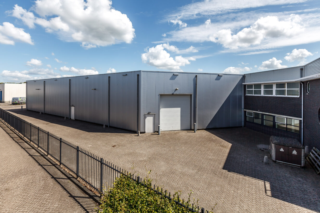 Groot industrieel pakhuis met metalen gevelbekleding en een gesloten witte garagedeur, grenzend aan een geplaveide binnenplaats en bakstenen gebouw onder een gedeeltelijk bewolkte hemel.