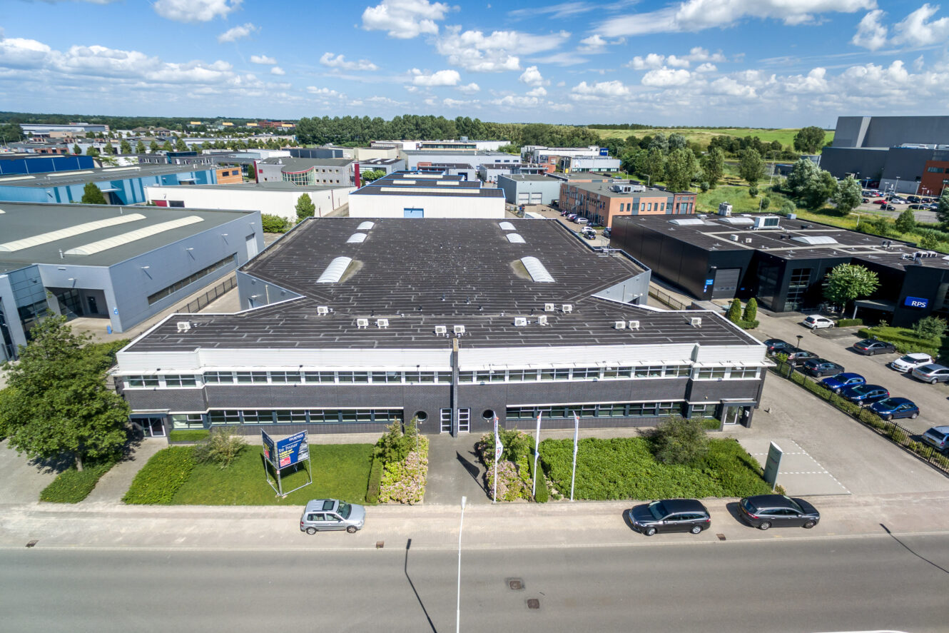 Een groot industrieel gebouw met een plat dak en veel ramen, omgeven door groen en geparkeerde auto's, gelegen op een bedrijventerrein met andere soortgelijke gebouwen.