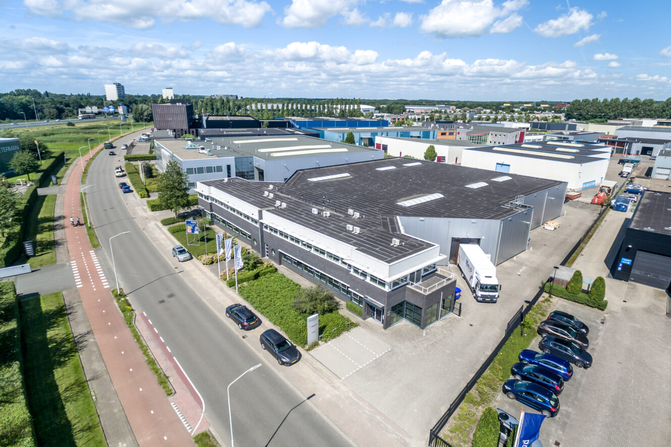 Luchtfoto van een industriegebied met grote magazijngebouwen, een parkeerplaats met auto's, een vrachtwagen bij een laadperron en een weg die langs het complex loopt.