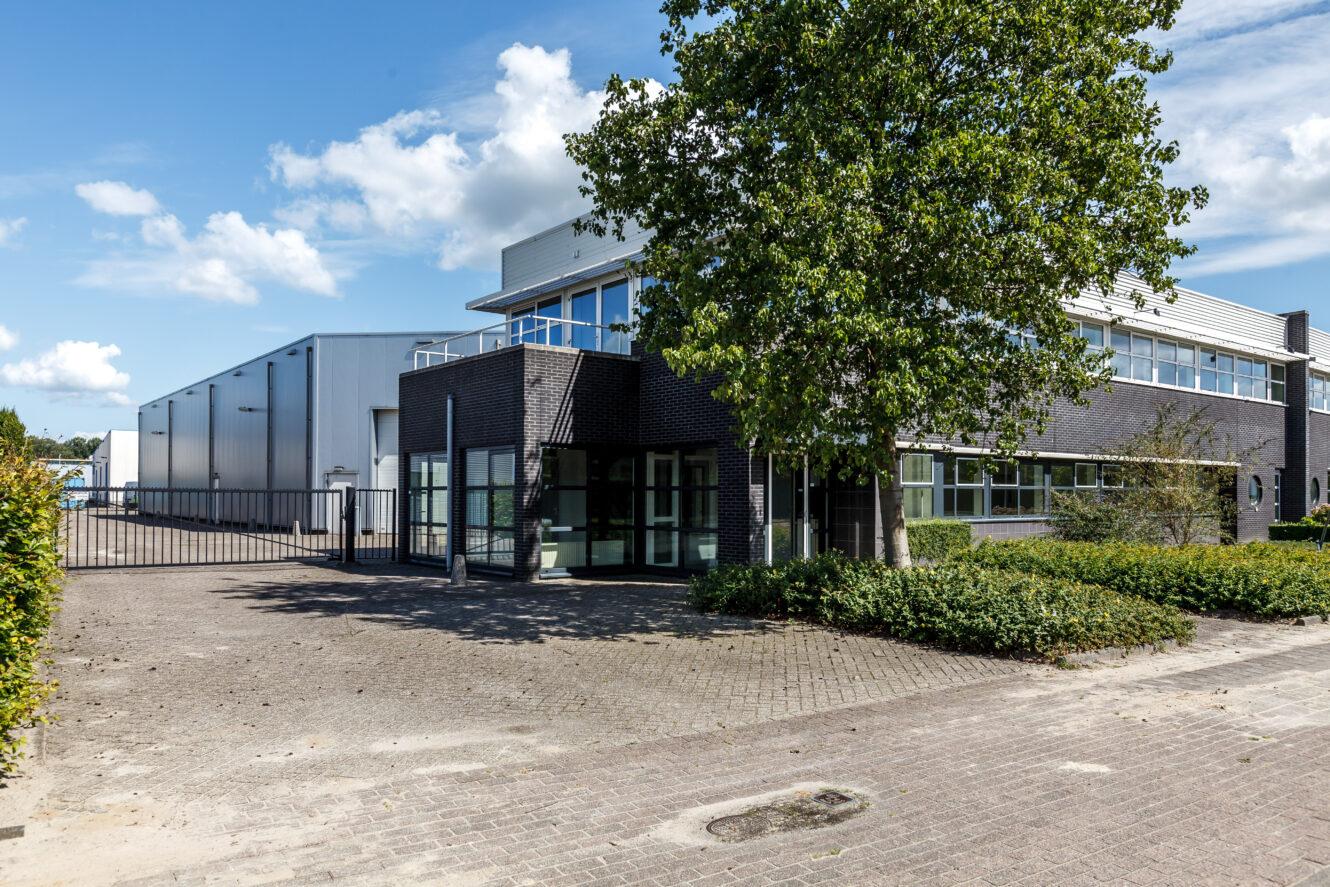 Modern industrieel gebouw met grote ramen, aangrenzend magazijn, poortingang en een boom ervoor, onder een blauwe lucht met verspreide wolken.