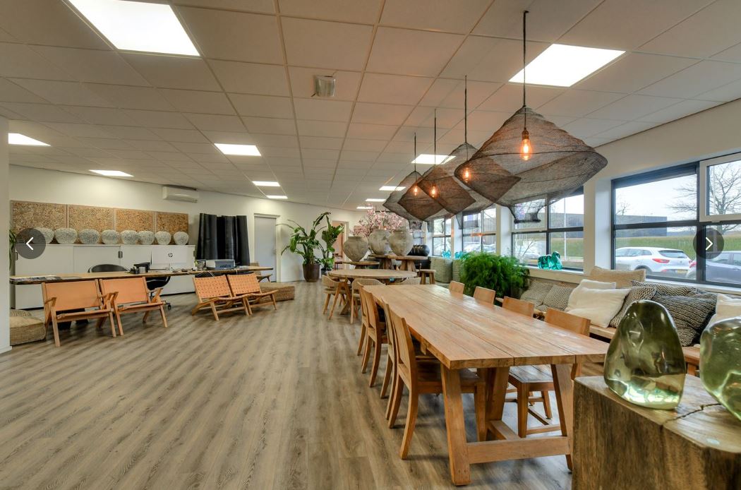 Een ruime kamer met houten tafels en stoelen, moderne verlichting, grote ramen en verschillende planten, met een zithoek en een kitchenette.