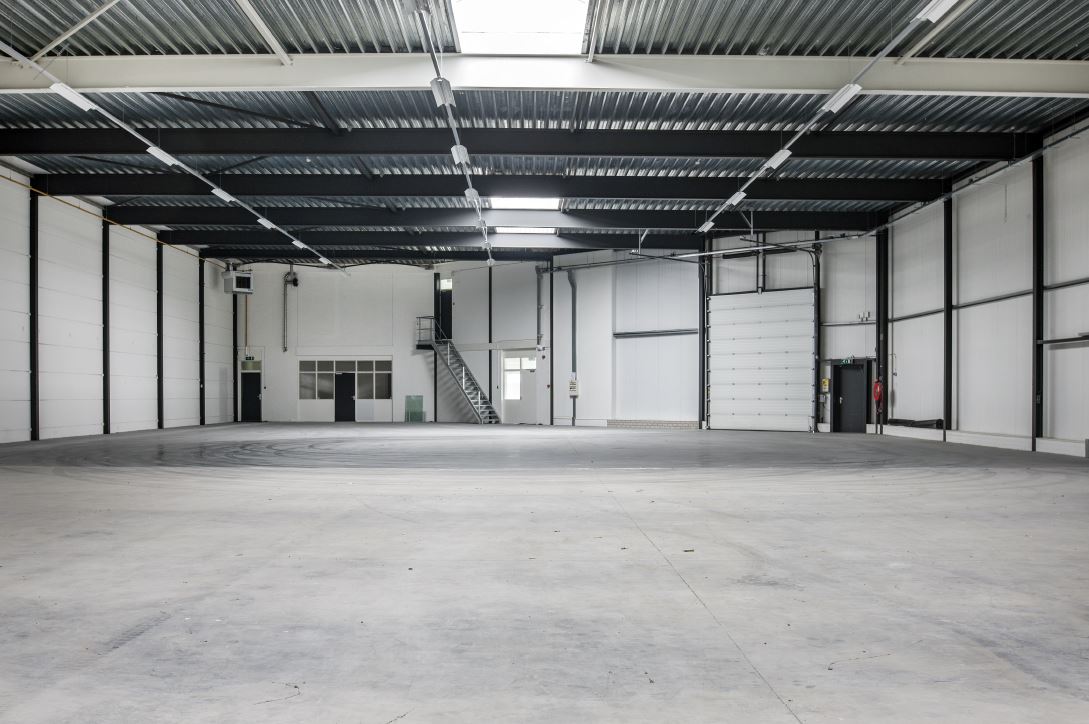 Een groot, leeg industrieel pakhuis met betonnen vloeren, witte muren, zwarte balken en een metalen trap naar een tussenverdieping.