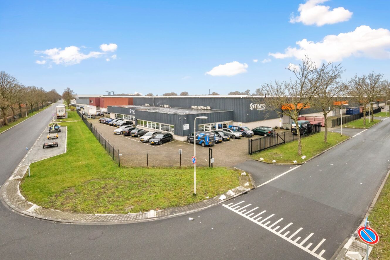 Industrieel gebouw met meerdere geparkeerde auto's op een omheind terrein, gelegen op een straathoek met omringend groen en een heldere hemel.