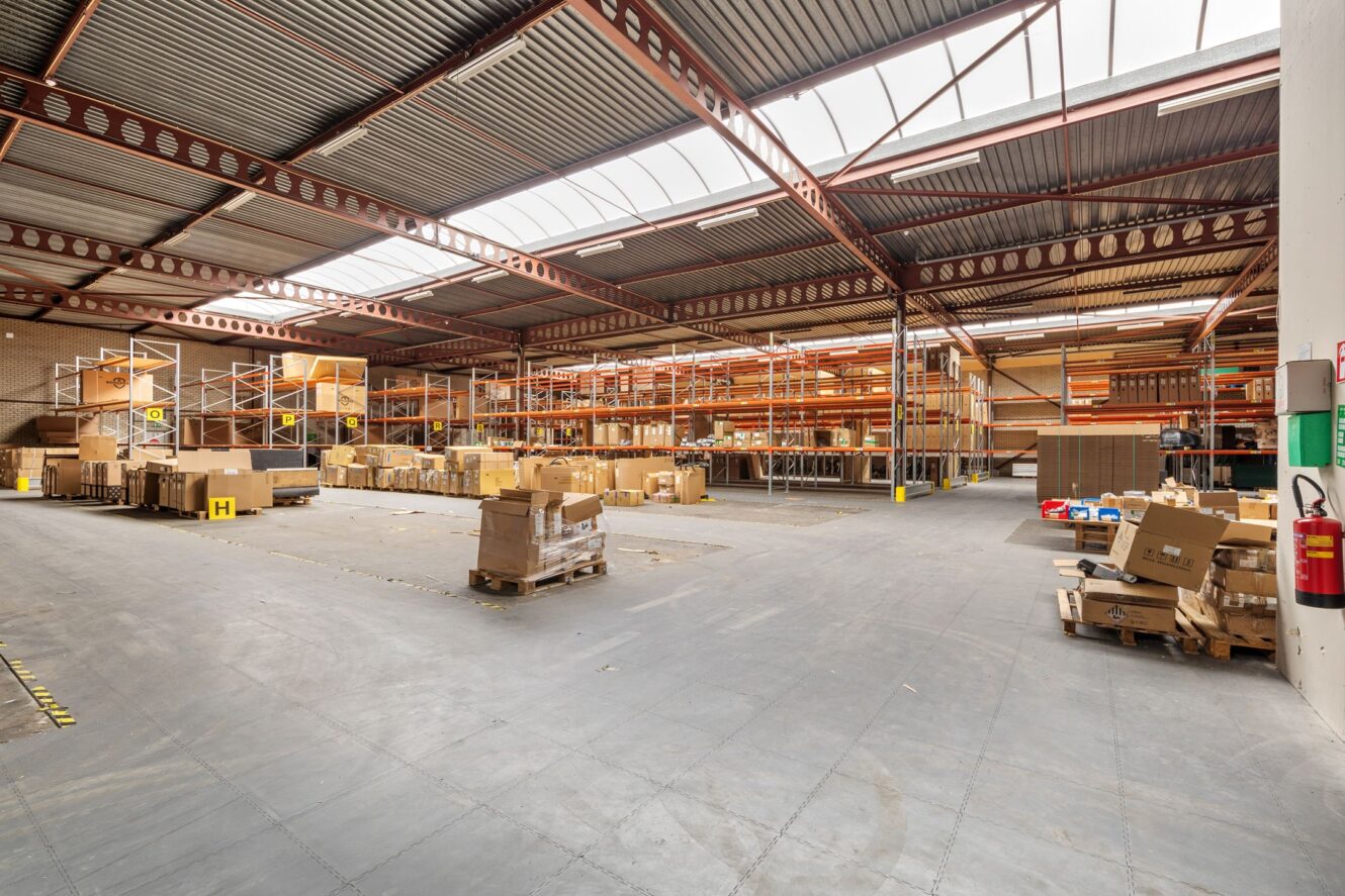 Groot magazijninterieur met metalen rekken, pallets met kartonnen dozen en open vloerruimte onder een hoog dak met lichtkoepel.