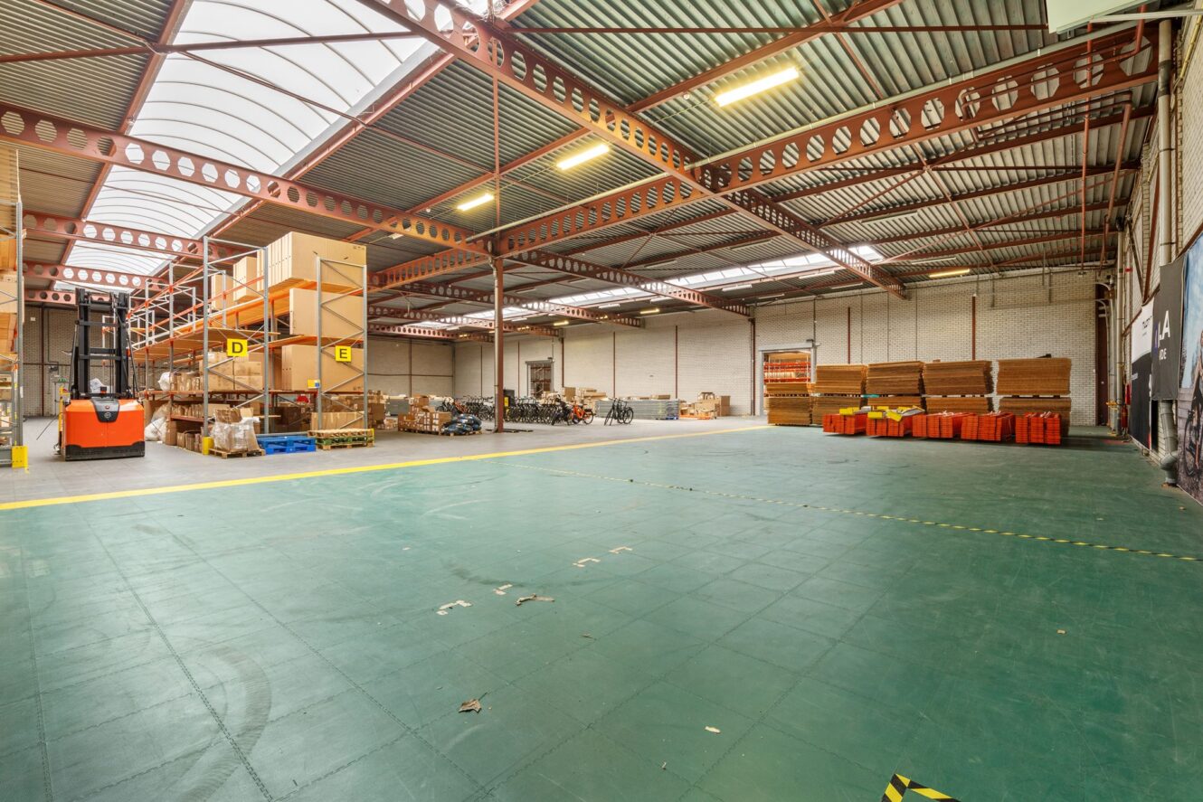 Groot magazijninterieur met hoge plafonds, industriële rekken, gestapelde materialen, een vorkheftruck en open vloerruimte gemarkeerd met gele lijnen.
