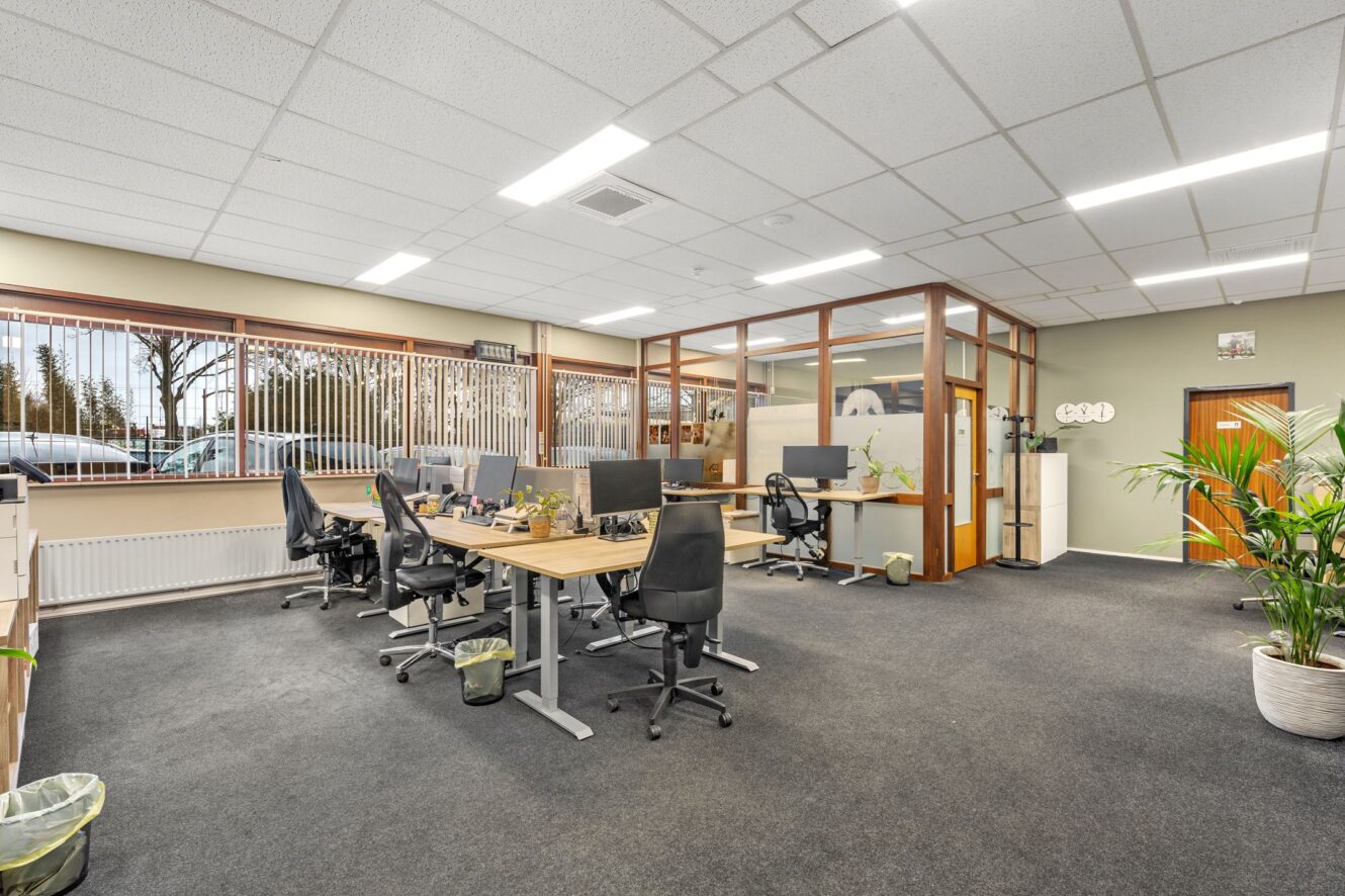 Modern open kantoor met verschillende werkplekken, zwarte stoelen, computers, kamerplanten, grote ramen en vergaderruimtes met glazen scheidingswanden. De ruimte is netjes en goed verlicht.