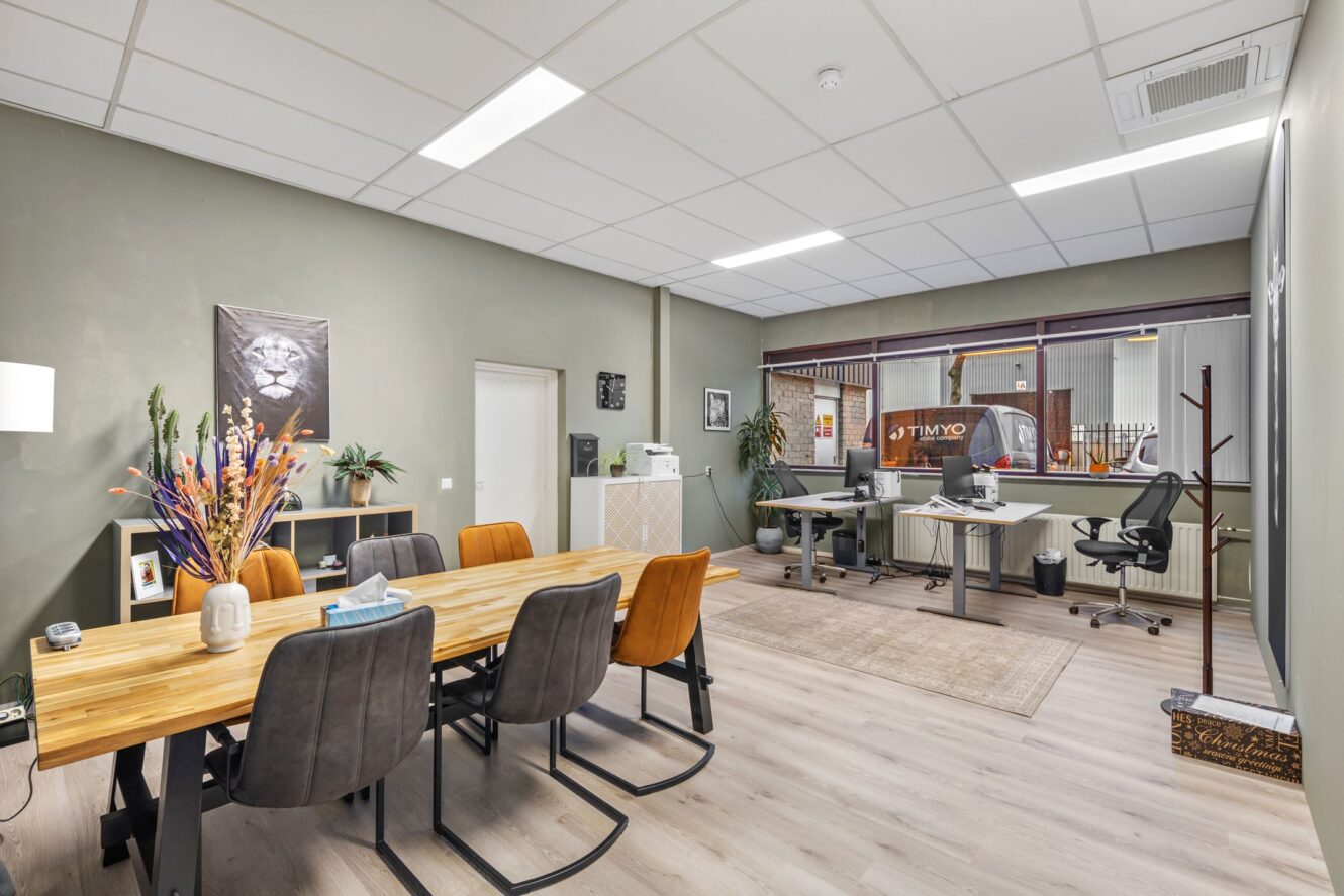 Modern kantoor met een houten vergadertafel, zes stoelen, bureaus met computers, neutraal gekleurde muren en een groot binnenraam dat een andere kantoorruimte laat zien.