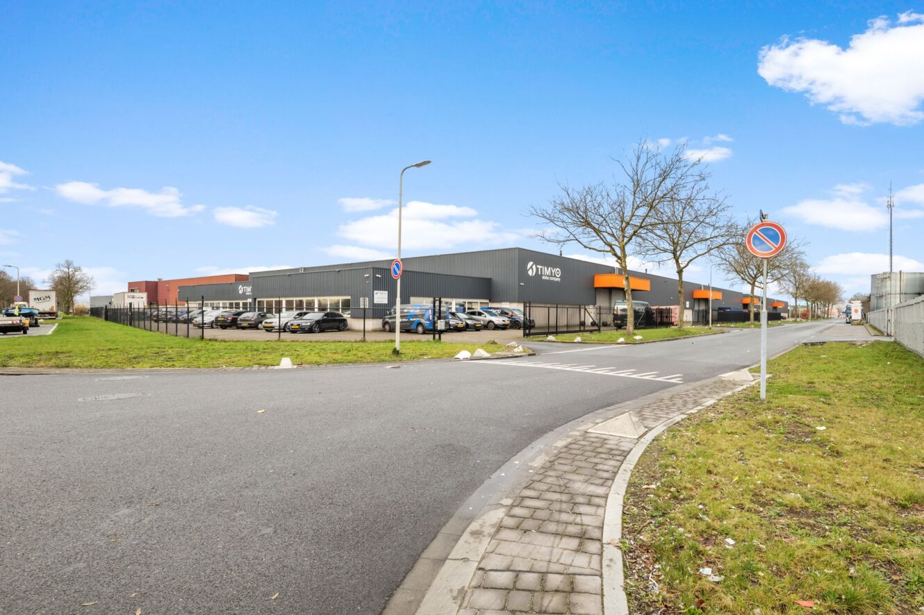 Groot magazijngebouw met TWWB-borden, een omheinde parkeerplaats met meerdere auto's, heldere hemel en verkeersborden met parkeerverbod langs een bochtige straat.