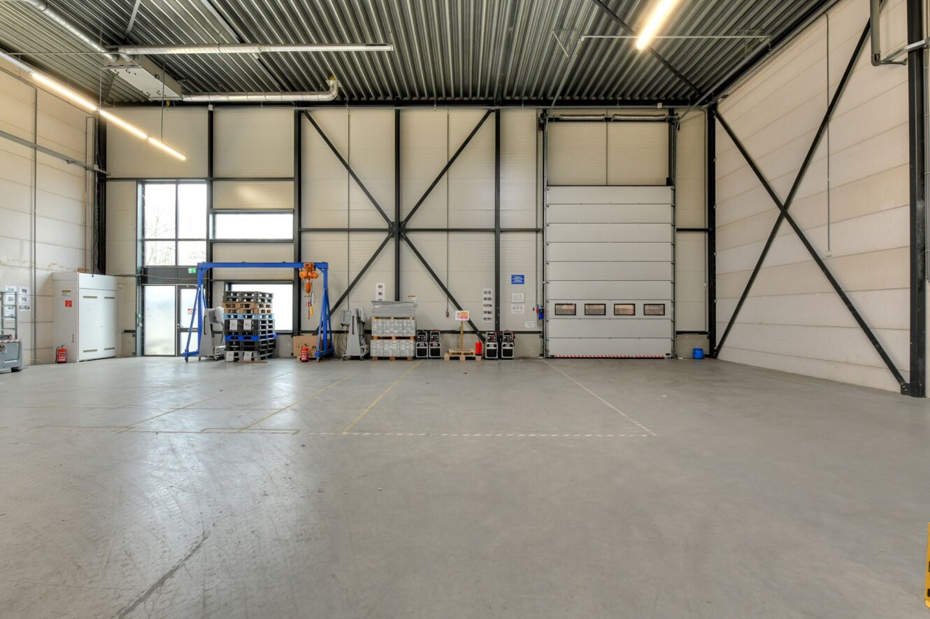 Ruim industrieel magazijninterieur met hoge plafonds, betonnen vloer, gestapelde pallets en machines langs de muren.