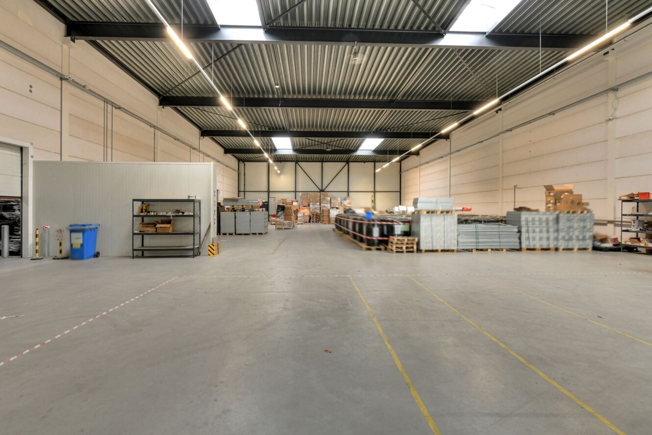 Groot magazijninterieur met hoge plafonds, industriële rekken en gestapelde pallets met dozen en materialen. De ruimte is schoon en goed verlicht.