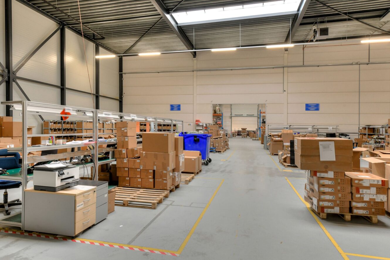 Groot magazijninterieur met hoge plafonds, rekken, werkstations en talloze gestapelde kartonnen dozen. Een printer en blauwe recyclingbakken zijn ook zichtbaar.