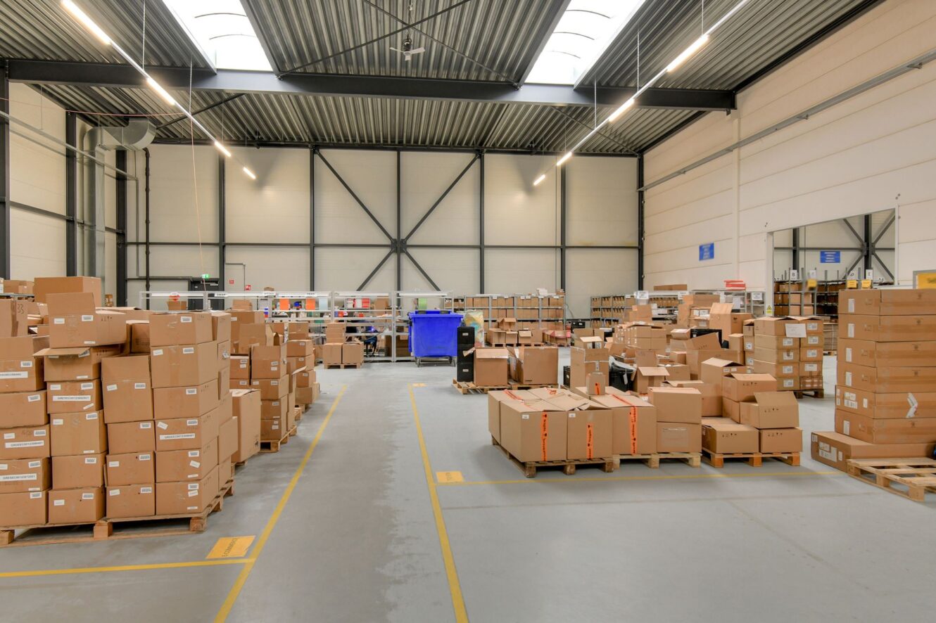 Een groot magazijninterieur met talloze kartonnen dozen gestapeld op pallets, georganiseerd in rijen onder heldere verlichting.