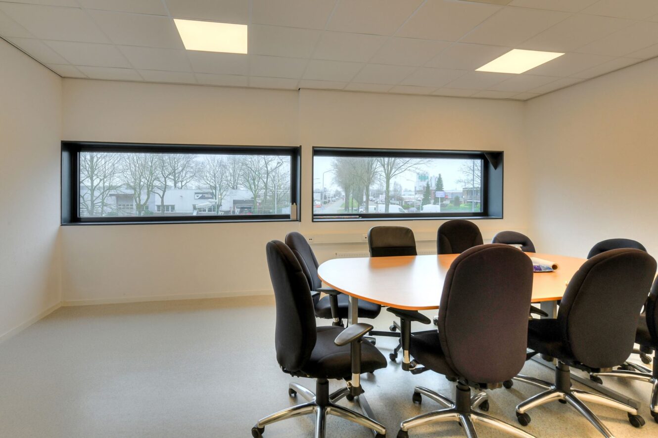 Een moderne vergaderruimte met een houten ovale tafel, acht zwarte bureaustoelen, grote ramen en uitzicht op bomen en gebouwen.