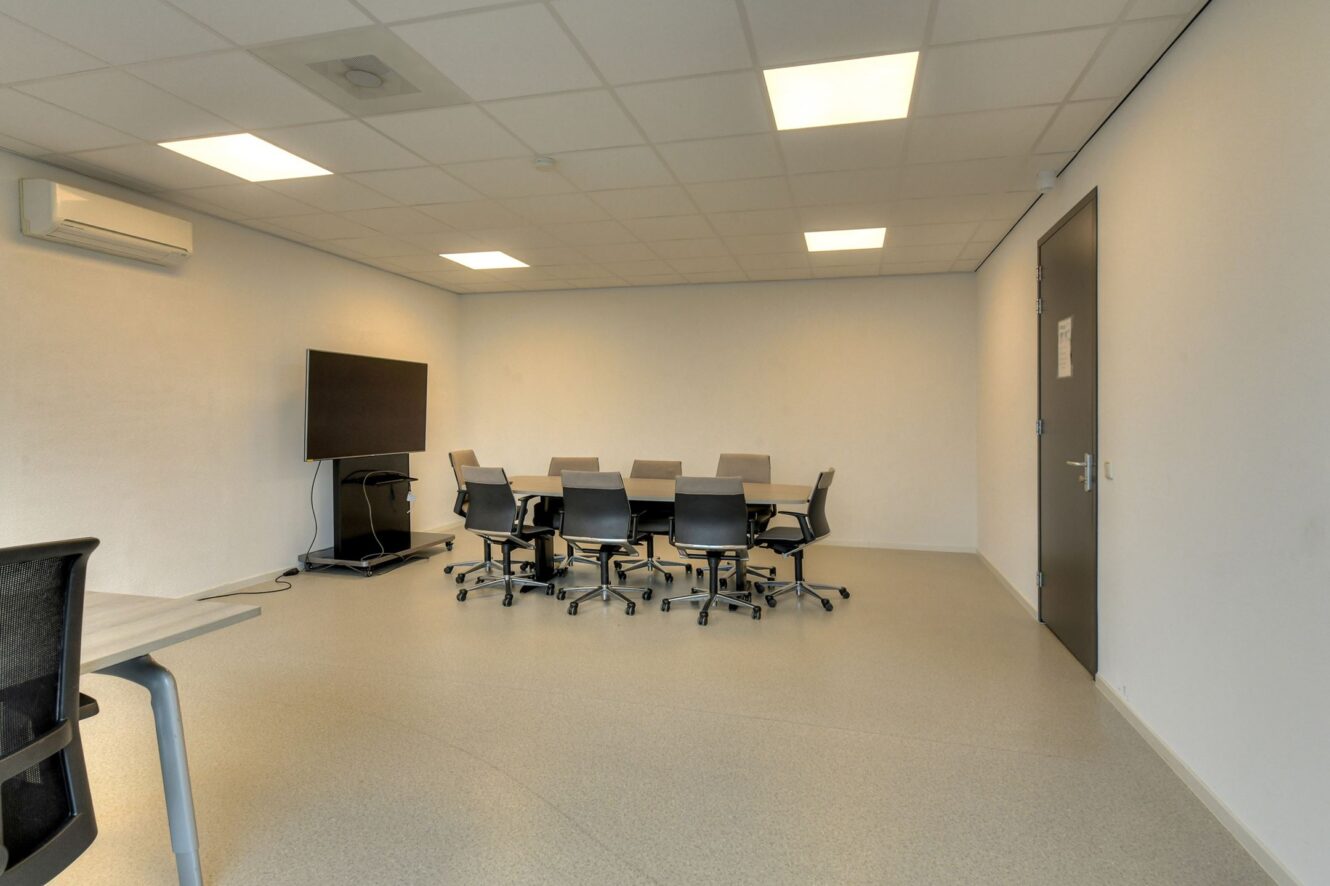 Een vergaderzaal met een tafel, zeven stoelen, een grote monitor op een standaard, een airconditioner en beige muren onder tl-verlichting.