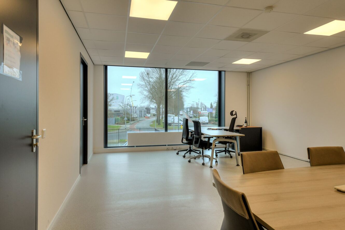 Modern kantoor met grote ramen, een bureau met computer, twee stoelen en een tafel met extra stoelen op de voorgrond. Buiten zijn bomen en een straat zichtbaar.