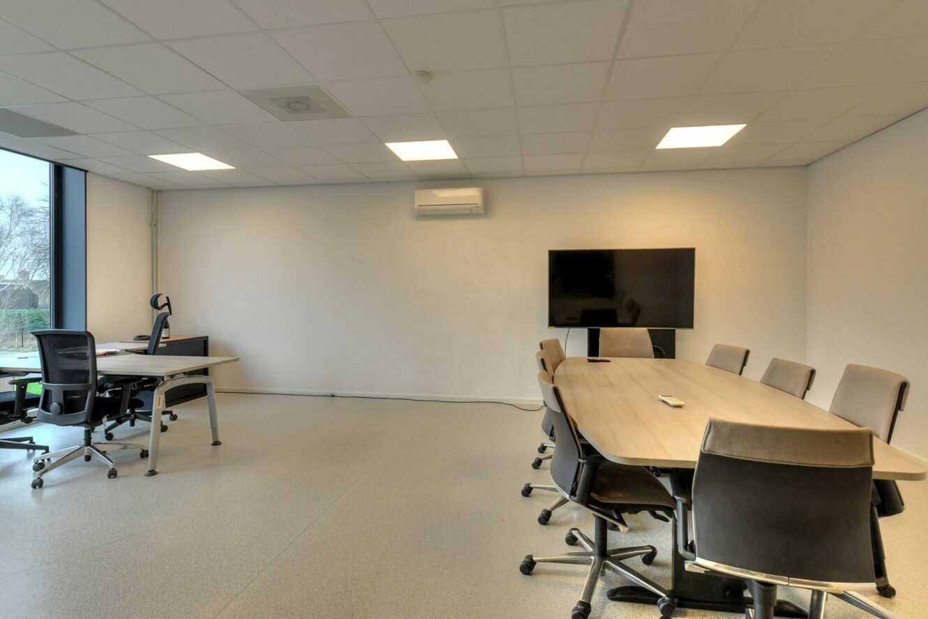 Moderne vergaderruimte met een lange tafel, acht stoelen, een groot scherm aan de muur en een apart bureau met een bureaustoel bij een raam.