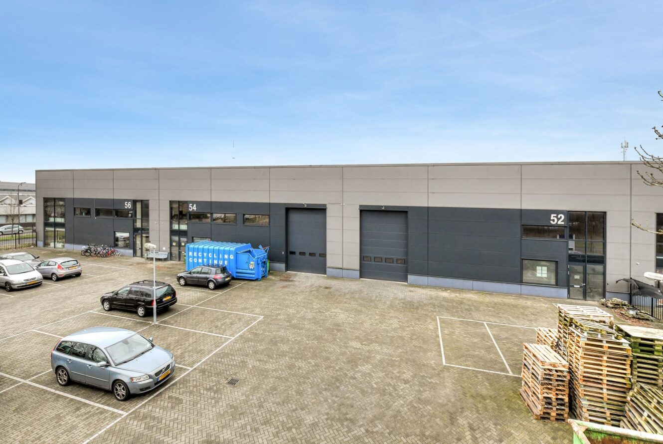 Parkeerterrein met verschillende auto's en een blauwe recyclingcontainer voor een modern industrieel magazijngebouw met drie laadperrons genummerd 52, 54 en 56.