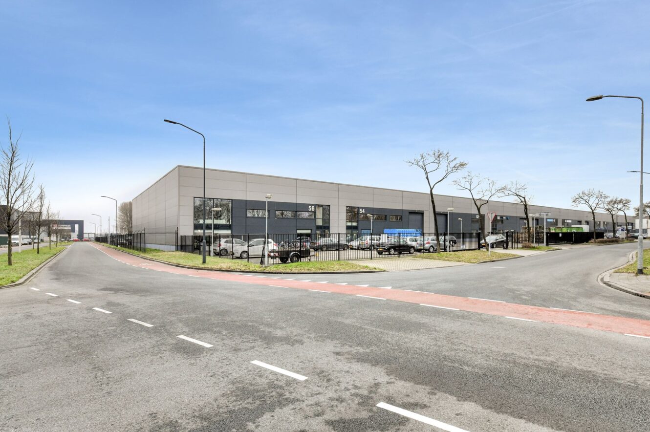 Een modern industrieel gebouw met grote ramen en laadkades, gelegen op het kruispunt van twee lege wegen onder een heldere hemel. Verschillende voertuigen staan buiten geparkeerd.