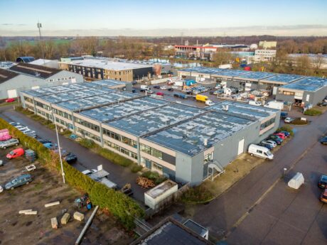Luchtfoto van een industrieterrein met meerdere magazijnen, geparkeerde voertuigen en aangrenzende wegen op een heldere dag.