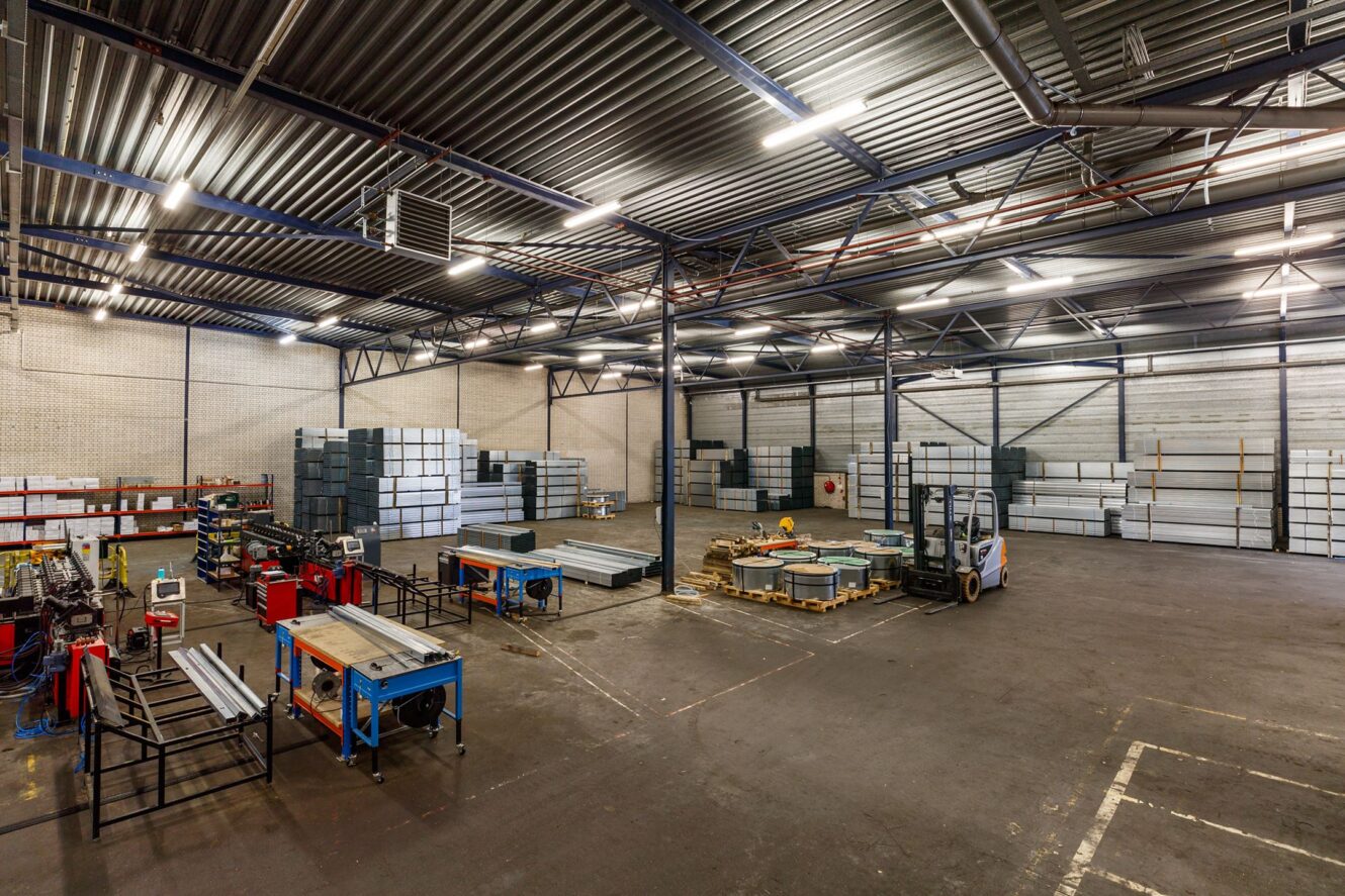 Interieur van een ruim magazijn met gestapelde metalen platen, rode en blauwe werkbanken en een vorkheftruck geparkeerd bij materialen op de vloer.
