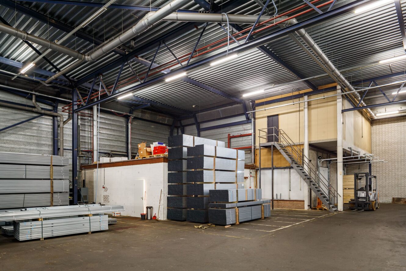 Interieur van een groot magazijn met stapels metalen balken, een vorkheftruck, industriële verlichting en een verhoogd kantoorgedeelte dat toegankelijk is via een trap.