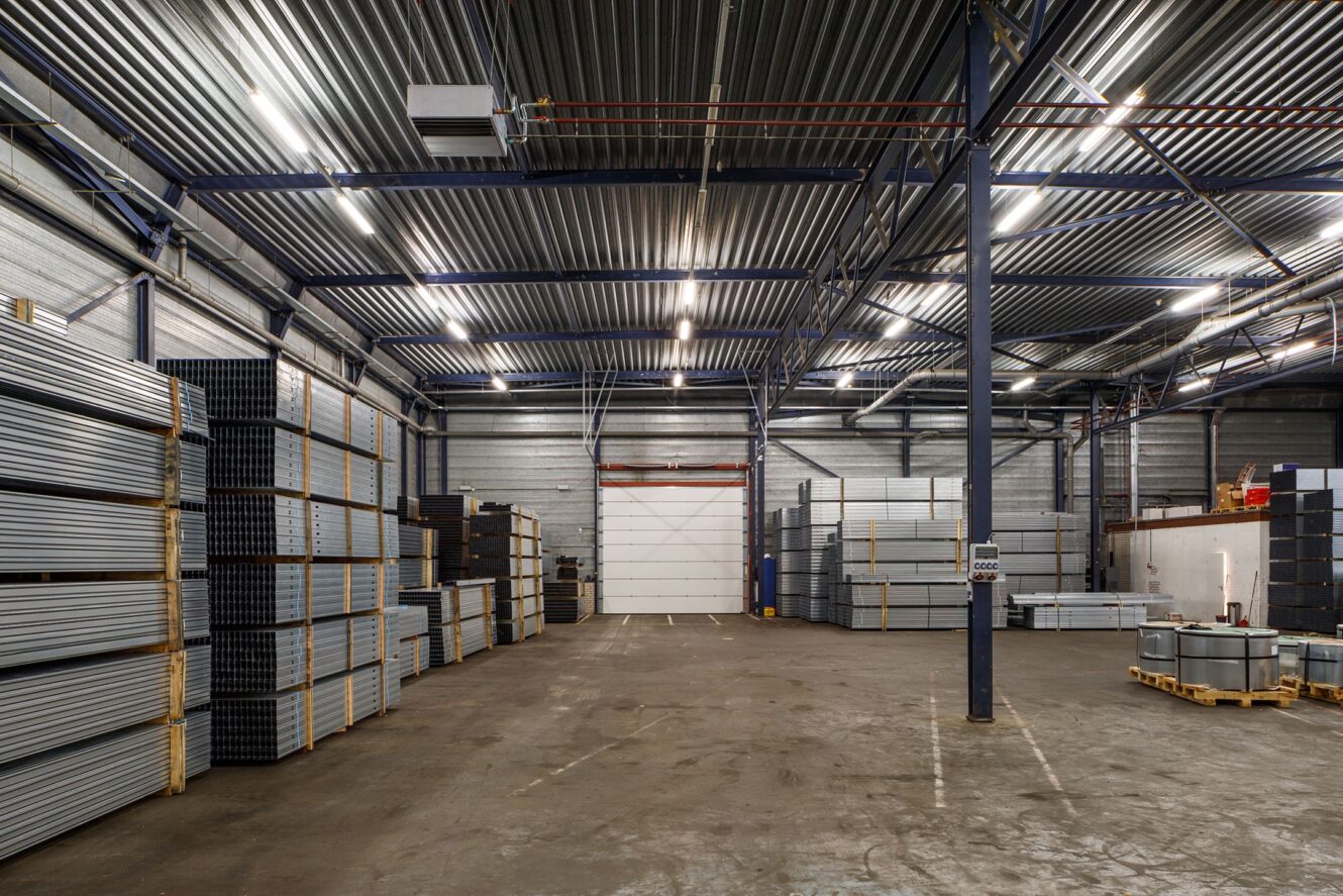 Interieur van een groot magazijn met gestapelde metalen buizen en platen, hoge plafonds, bovenverlichting en een gesloten witte garagedeur aan het einde.