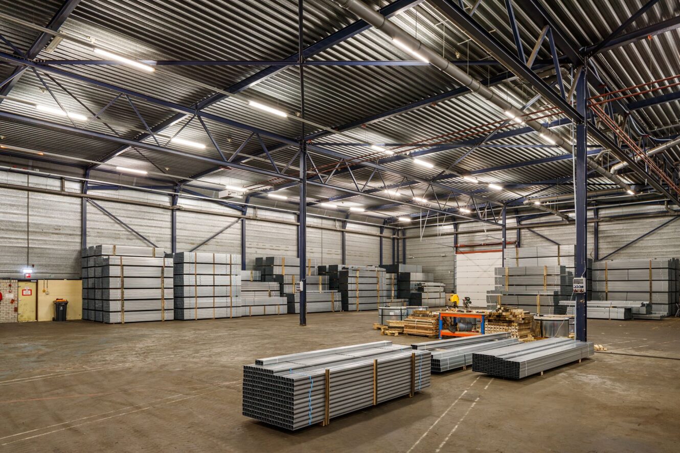 Interieur van een ruim magazijn met stapels metalen platen en balken langs de muren en wat materialen op houten pallets in het midden.