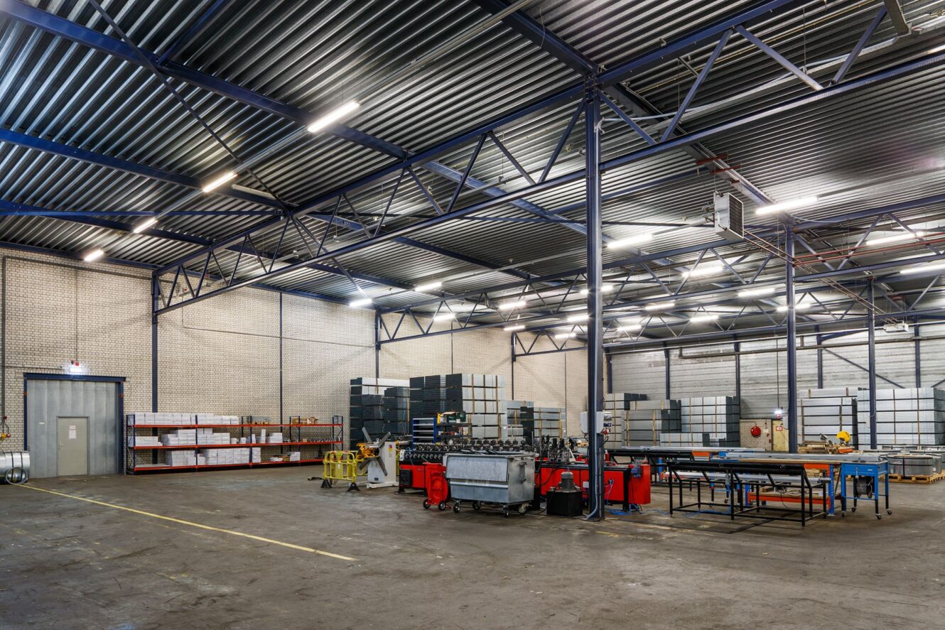 Ruim magazijninterieur met hoge plafonds, industriële verlichting, gestapelde pallets, metalen rekken en werkstations verspreid over de ruimte.