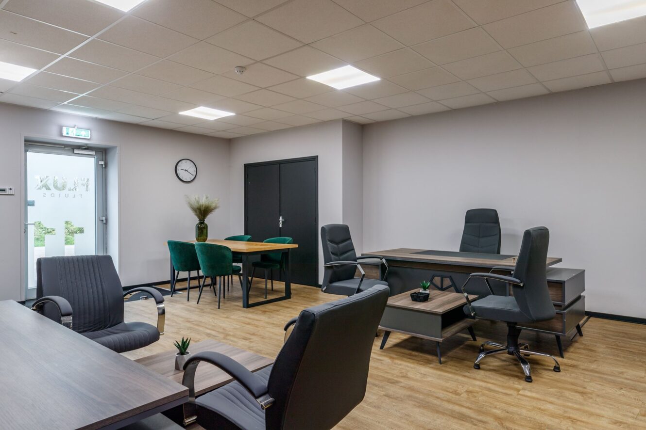 Modern kantoor met houten vloeren, verschillende bureaus en stoelen, een tafel met vier groene stoelen, een wandklok en een glazen deur met daglicht.