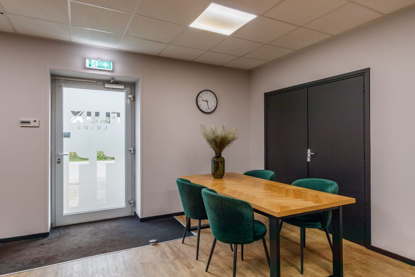 Een kleine vergaderruimte met een houten tafel, vier groene stoelen, een vaas met gedroogde planten, een wandklok, dubbele deuren en een glazen uitgang met matte letters.
