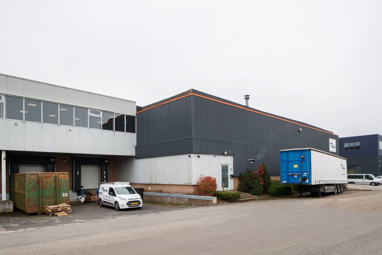 Industrieel magazijn aan de buitenkant met een laadperron, geparkeerde witte bestelbus, groene bakken, blauwe trailer en minimale tuinaanleg op een bewolkte dag.