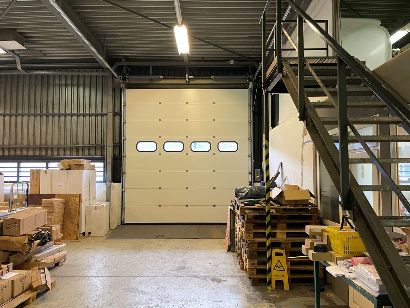 Magazijninterieur met een gesloten garagedeur, gestapelde dozen en pallets op de vloer en een metalen trap naar een bovenverdieping.
