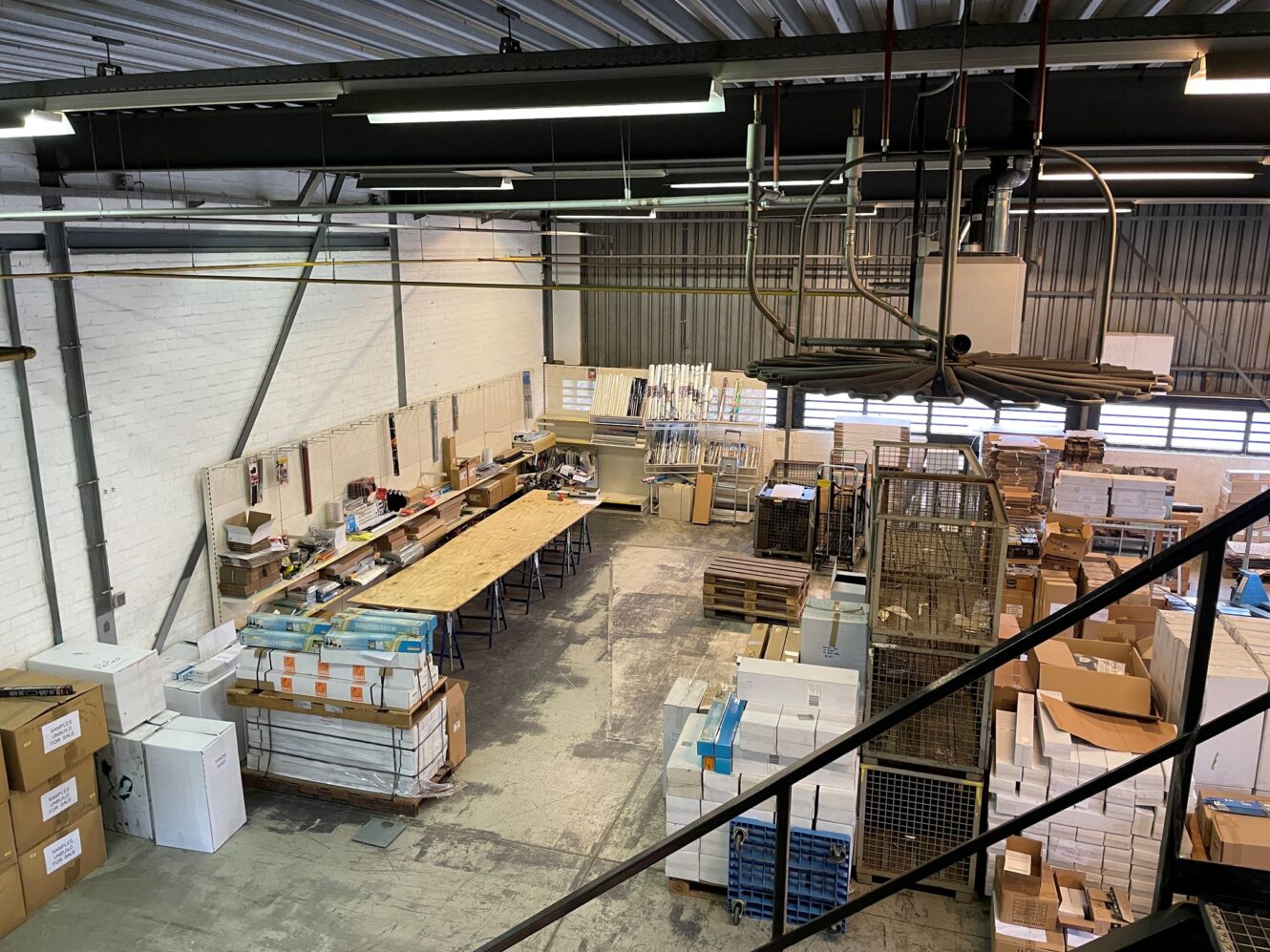 Een magazijnwerkplek met lange tafels, planken, gestapelde dozen en diverse apparatuur, gezien vanuit een verhoogd perspectief.