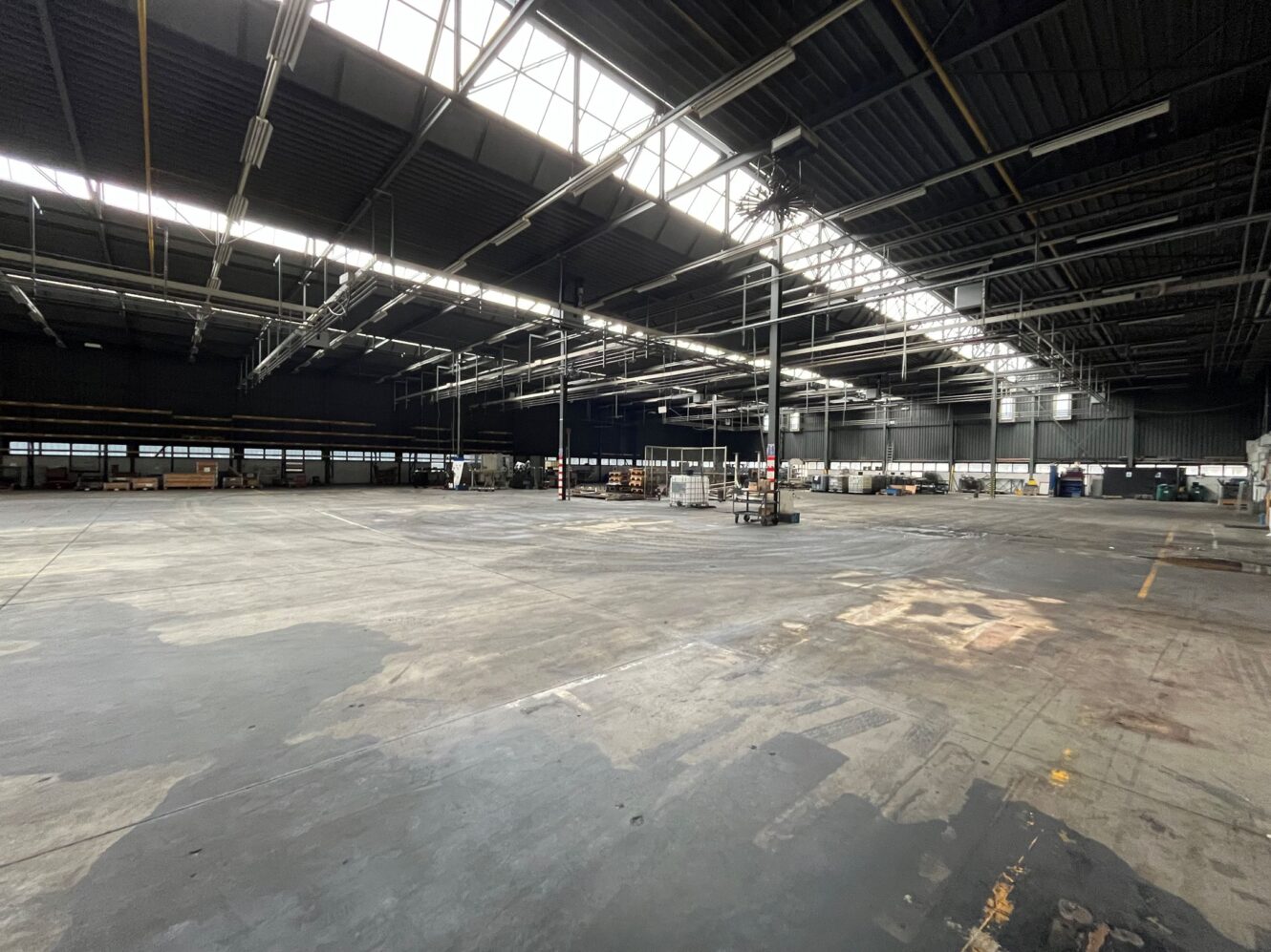 Groot, grotendeels leeg magazijninterieur met hoge plafonds, zichtbare balken, dakramen en verspreide industriële apparatuur langs de omtrek.