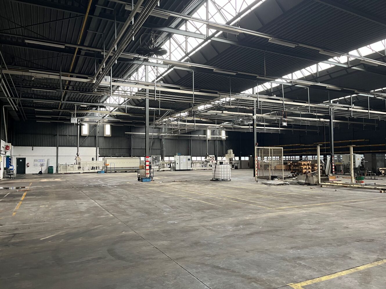 Ruim, meestal leeg industrieel pakhuis met hoge plafonds, dakramen en verspreide apparatuur en materialen langs de muren.